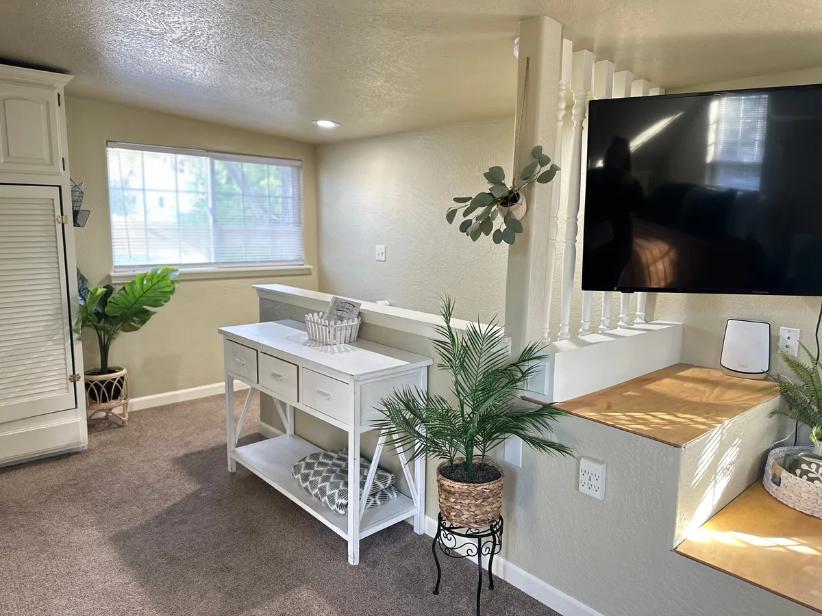 A bright corner featuring a white console table topped with potted plants, a TV mounted on the wall, and a window letting in natural sunlight. The overall mood is cozy and welcoming, ideal for relaxing or enjoying entertainment.