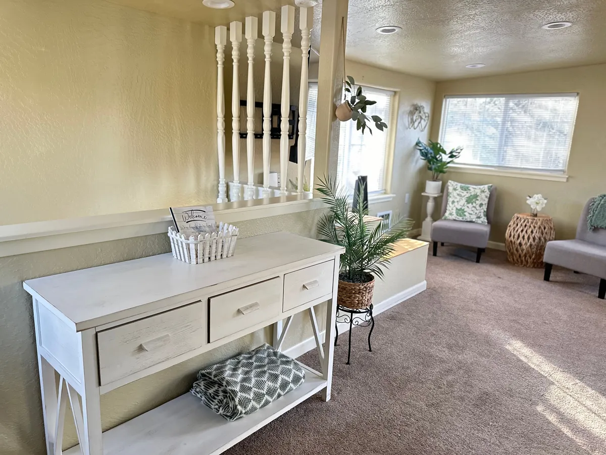 A cozy seating area featuring a white console table adorned with green plants, plush decorative pillows, and comfortable seating. The setup receives ample natural light and offers a welcoming environment ideal for unwinding or enjoying a cup of coffee.