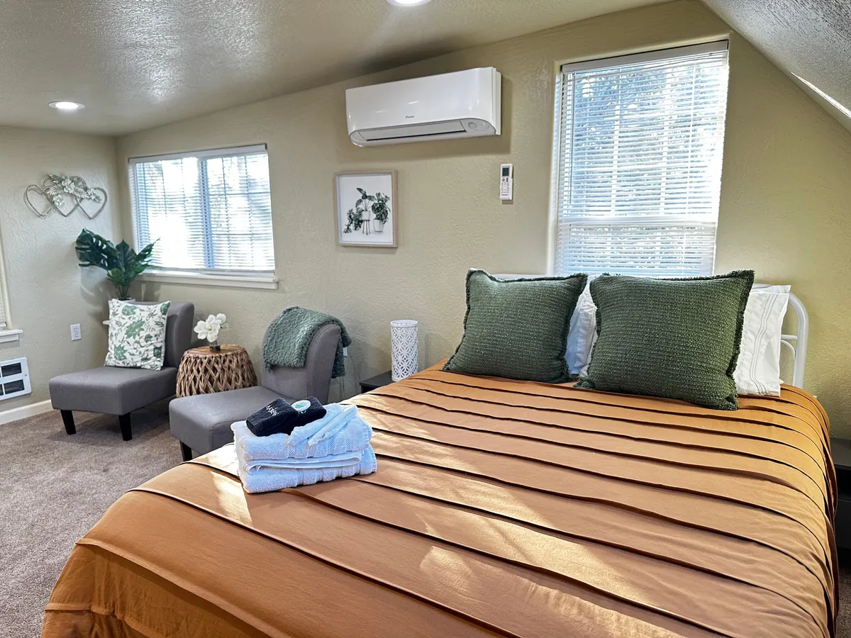 The image features a bright, welcoming studio interior with a plush bed dressed in textured orange bedding and green accent pillows. Sunlight streams in, illuminating a cozy seating area perfect for relaxing with a book or morning coffee. The overall atmosphere is warm, stylish, and inviting, hinting at a peaceful retreat close to downtown Medford.