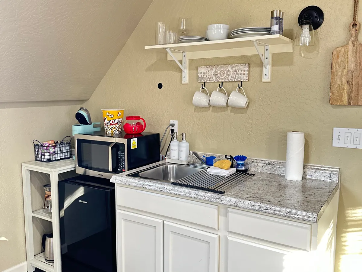 A close view of a kitchen sink set beneath open shelves holding dishes, mugs, and a microwave. Coffee mugs are prominently displayed, and the overall atmosphere is warm and homey, perfect for casual meal prep or coffee breaks.