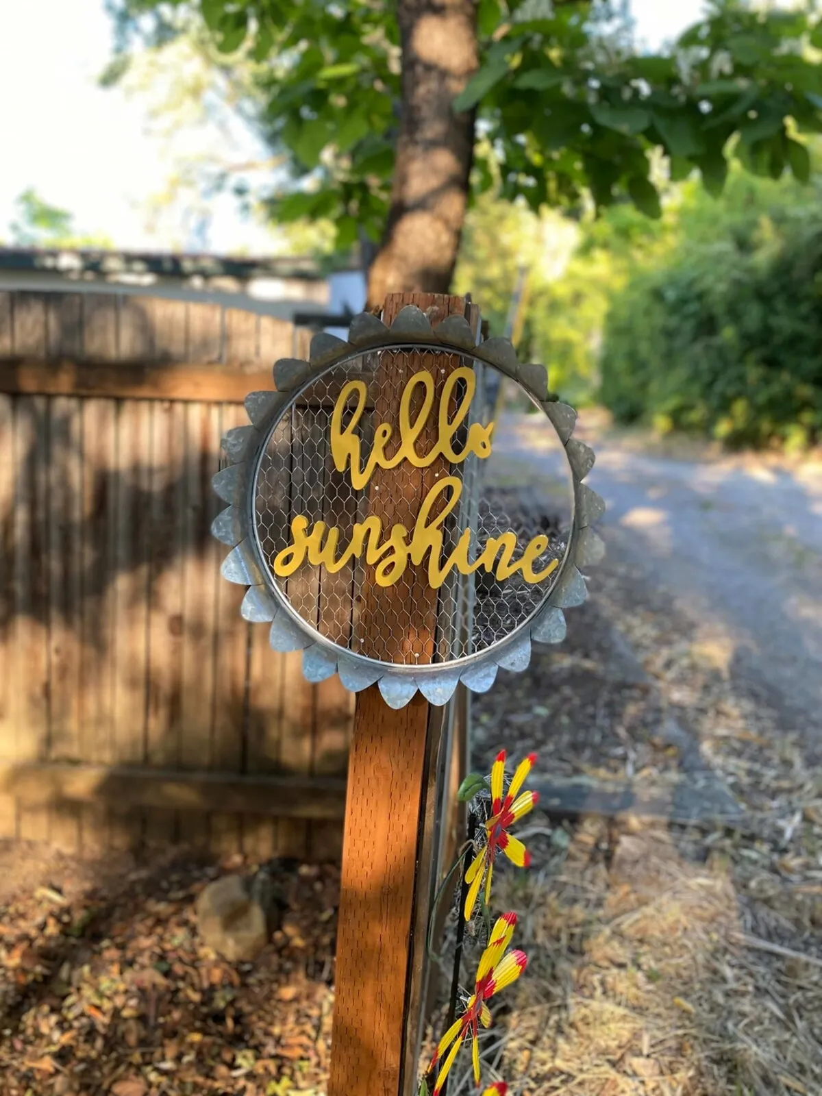 An exterior scene featuring a decorative sign surrounded by lush natural greenery and colorful embellishments. The garden pathway is inviting, suggesting a welcoming entrance and easy outdoor access, with hints of neighborhood life just beyond.