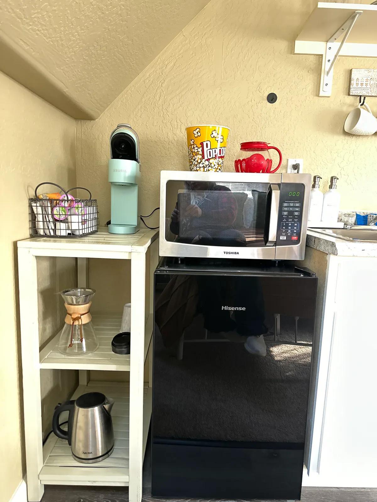 A compact kitchenette featuring a microwave, mini fridge, coffee maker, and open shelving. The countertop is organized for easy access to meal and beverage prep, with a popcorn container and coffee supplies adding a sense of comfort and convenience.